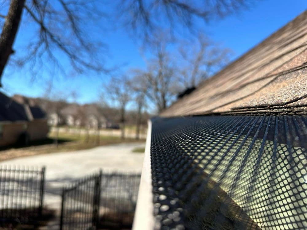 Close-up of a roof edge with mesh gutter guard, trees and a clear blue sky in the background.