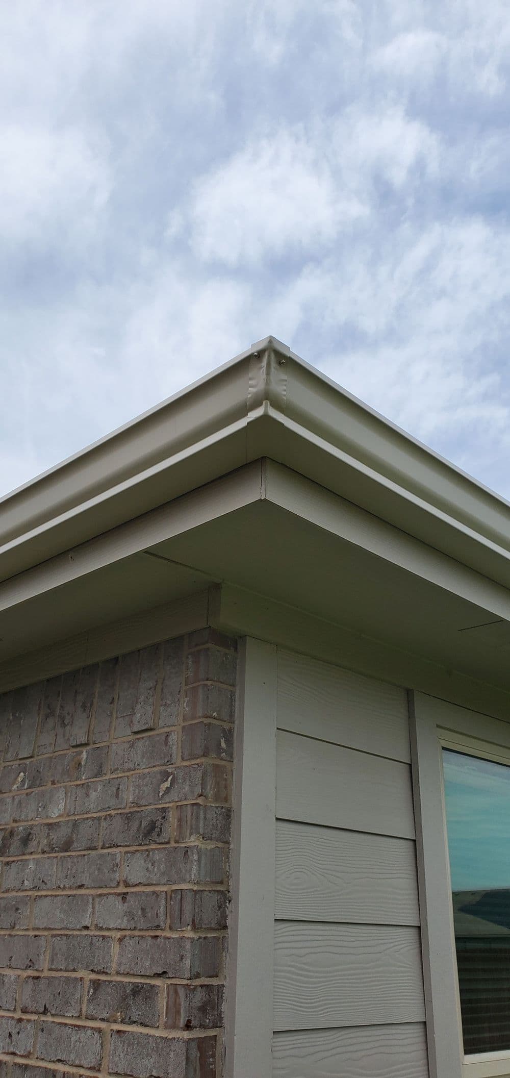 Roof edge and corner of a house with brown brick and light-colored siding under a cloudy sky.