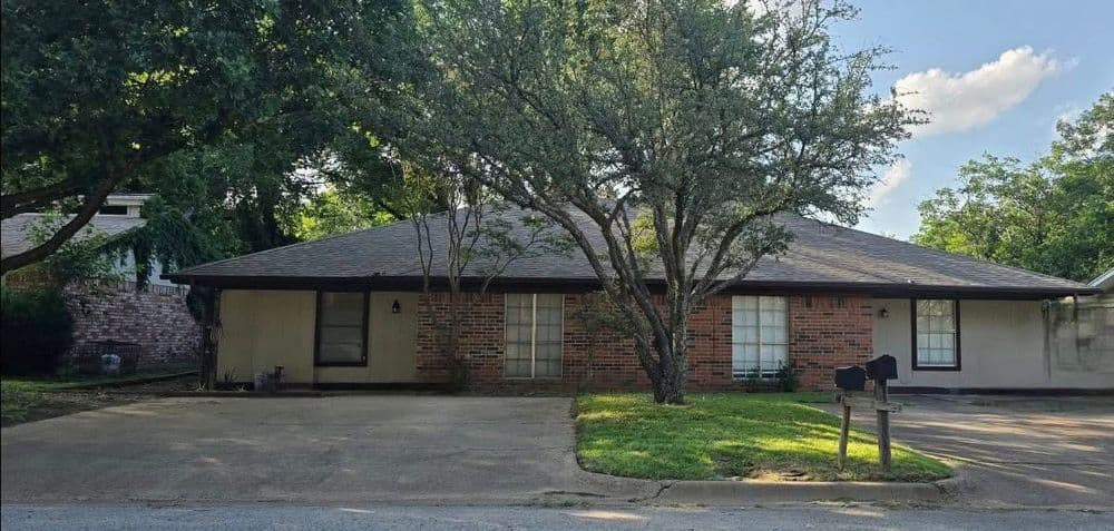 Single-story brick house with large front yard, shaded by a mature tree and driveway.