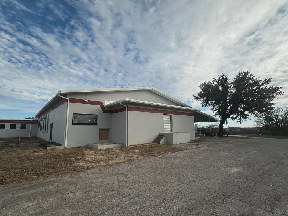 Modern gray building with red accents, surrounded by open land and a tree under a cloudy sky.