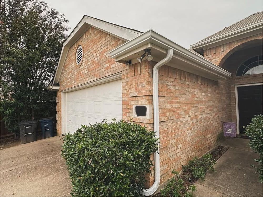 Brick house exterior with garage, landscaping, and trash bins visible on the driveway.